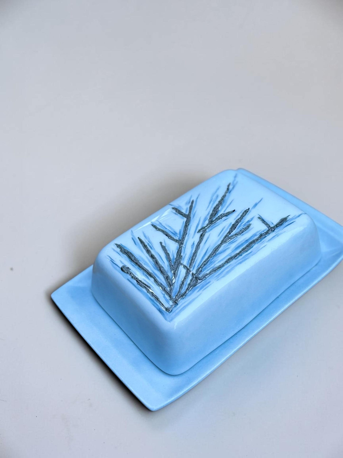 May include: A light blue ceramic butter dish with a rectangular base and a matching lid. The top of the lid features a dark grey, textured, hand-painted branch or twig design that contrasts with the smooth, matte finish of the light blue glaze.